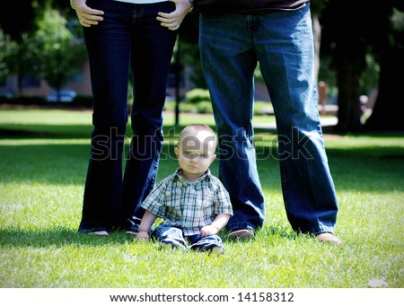 A small child seated in the grass, the parents are standing above him. - vertically framed