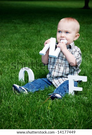 A baby sitting in a field of grass eating a letter N. - vertically framed