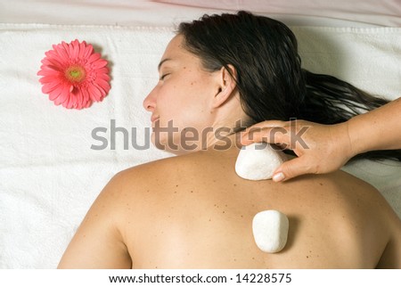 A girl getting a white rock placed on her back. A pink flower is near her as she lies on her white towel. - horizontally framed