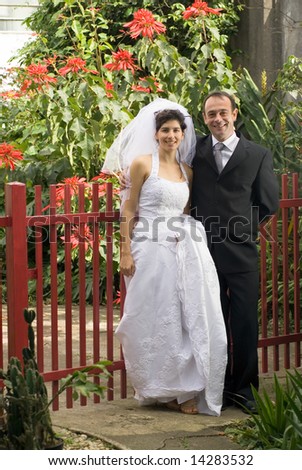 Married couple pose in front of a red fence, with red flowers in background. - vertically framed