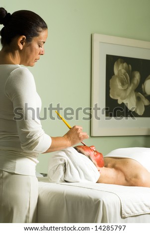 Woman lies down as another woman applies a red face mask with a paintbrush. Vertically framed photograph
