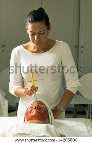A relaxed smiling woman getting a red facial mask brushed on at a spa.  Vertically framed shot.