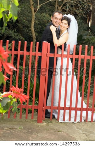 A couple, newly married, smile for the camera while hugging behind a red gate. - vertically framed