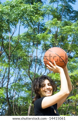 A smiling woman is playing basketball on a court at the park.  The woman is looking at the camera and about to make a shot.  Vertically framed photo.