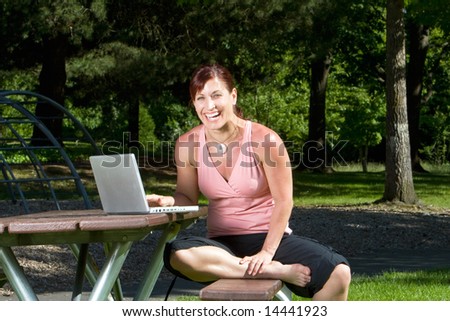 Woman laughs as she works on her laptop on a park bench. Vertically framed photograph