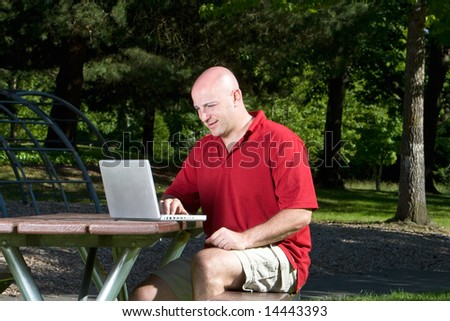 Man smiles as he works on his laptop on a park bench. Horizontally framed photograph