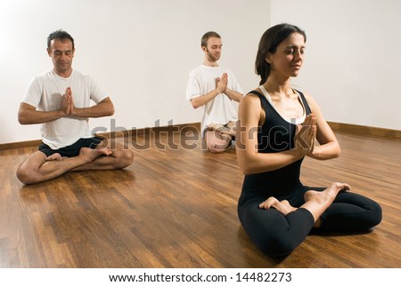 Two men and a woman practice yoga in a studio, they are sitting cross legged with their hands together and their eyes closed. Horizontally framed photograph