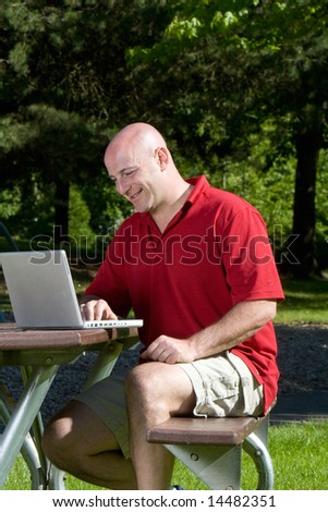 Man smiles as he works on his laptop on a park bench. Vertically framed photograph