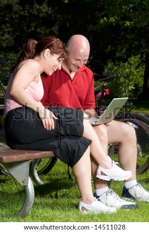 Couple on a park bench laugh as they look at a laptop computer, their bikes are in the background. Vertically framed photograph