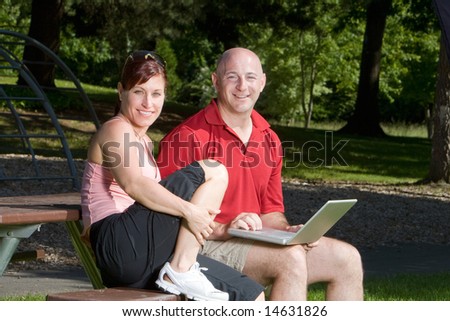 Couple lovingly sitting on a picnic table smiling and sitting at the park. The man is holding a laptop computer. Horizontally framed shot.
