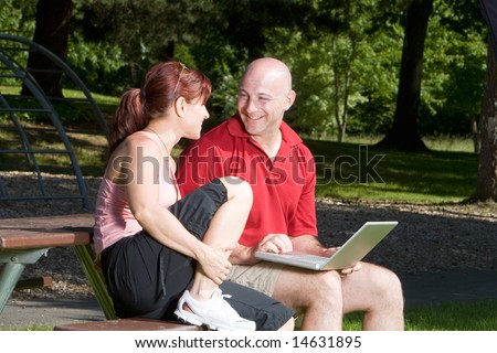 Couple lovingly sitting on a picnic table smiling and looking at each other in the park. The man is holding a laptop computer. Horizontally framed shot.