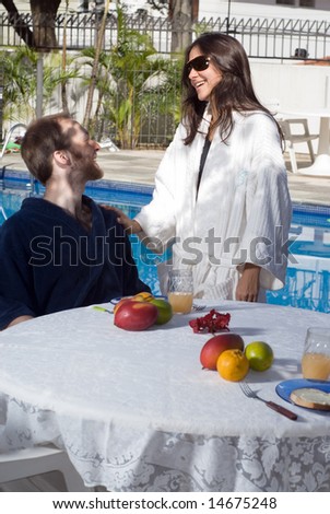 A young, attractive couple, the man sits down in a black robe, at a fruit covered table, the woman stands next to him in a white robe. - vertically framed