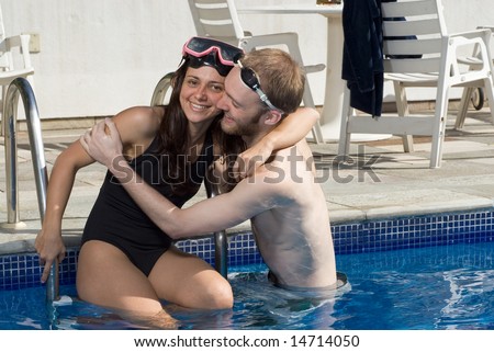 A man and woman are standing together in a pool.  They are hugging each other.  The man is looking at the woman, and the woman is looking at a camera.   Horizontally framed photo.