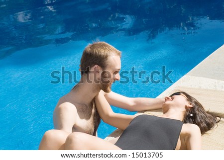 A young couple near the pool. A young woman, lies on the pools edge, while a young man stands in the pool\'s water, both staring and touching each other. - horizontally framed