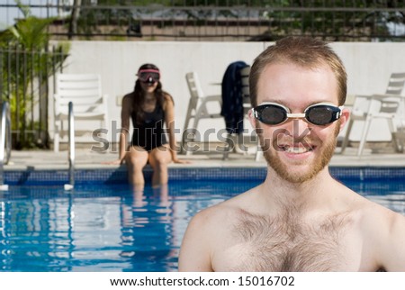 A man and a woman are sitting in a pool.  The woman is on the far end of the pool looking at the camera.  The man is standing right in front of the camera looking at it.  Horizontally framed photo.