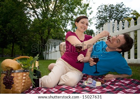 A couple toasting wine glasses on a picnic spread.  They are joking with each other and she is feeding him grapes. Horizontally framed shot.