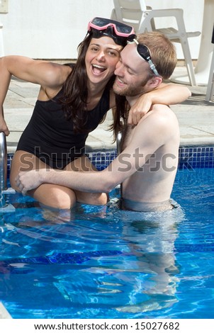 A man and woman are standing together in a pool.  They are hugging each other and laughing.  The man is looking at the woman, and the woman is looking at a camera.   Vertically framed photo.