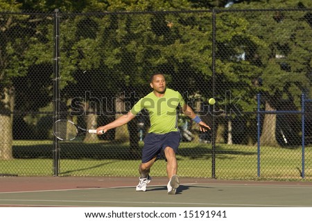 A man is outside on a tennis court playing tennis.  His is about to swing and is looking at the tennis ball.  Horizontally framed shot.
