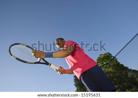 A man is outside on a tennis court playing tennis.  He is just about to throw the ball up to serve it and looking away from the camera.  Horizontally framed shot.