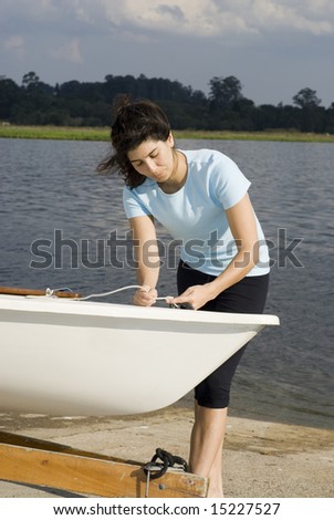 A woman is standing next to a sailboat on a lake.  She is tying a knot and looking at the rope.  Vertically framed photo.