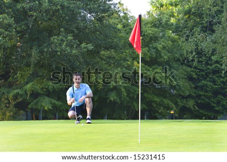 A man kneels, as he tries to line up a shot while golfing. Horizontally framed shot.