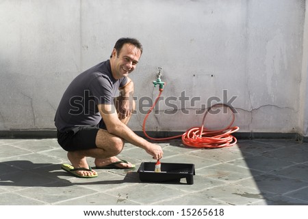 Man squatting over paint tray with hose in background. Man wearing shorts and sandals. Horizontally framed photo.