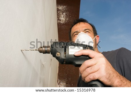 A man is holding a power drill and drilling into a wall.  He is wearing a mask and looking away from the camera and the drill.  Horizontally framed shot.