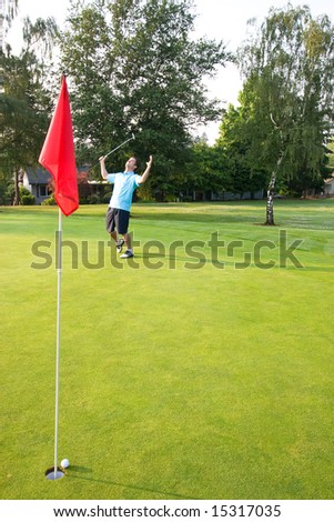 A man is standing on a golf course.  He just putted the ball into the hole.  His arms are raised and he is looking at the flag.  Vertically framed shot.