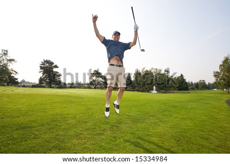 A man is jumping up and down on a golf course.  He is holding a golf club and smiling at the camera.  Horizontally framed shot.