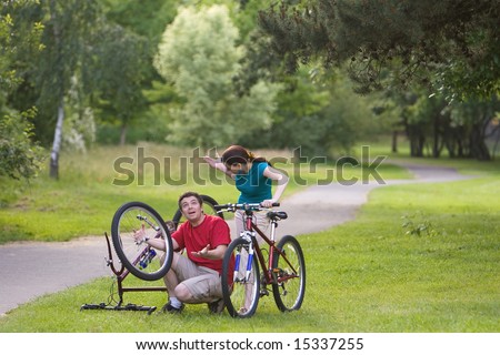 A man, sitting down, attempts to fix his bicycle, while it is upside-down and explaining it to the woman. The woman stands behind him, watching. - horizontally framed