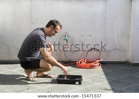 Man kneeling while dipping paint brush in paint tray. He is looking at paint tray and smiling. Horizontally framed photo.
