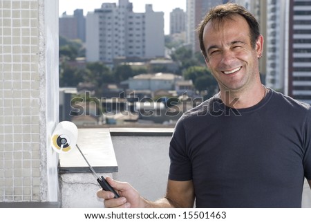A man is painting an unfinished apartment.  He is holding a paint roller and smiling.  Horizontally framed shot.