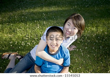 A young child is sitting on his mom's lap in a park.  They are smiling and looking at the camera.  Horizontally framed shot.