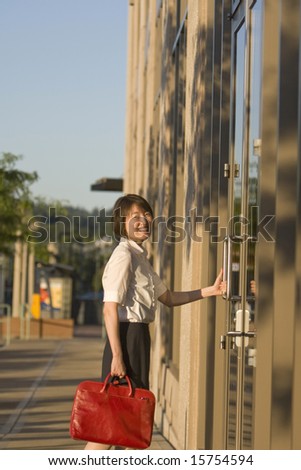 Young woman smiles at camera while opening a door to a building. She is smiling at the camera and carrying a red bag. Vertically framed photo.
