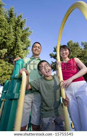 Smiling family stands on jungle gym looking at camera. Boy stands in front making a face at the camera. Vertically framed photo.
