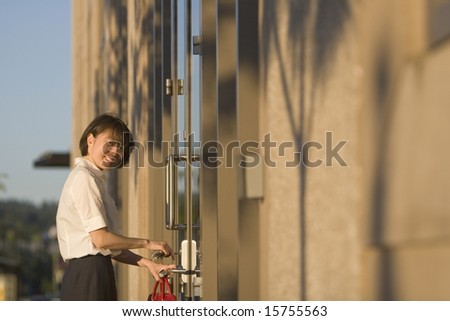 Young woman opens door while smiling at the camera. She is holding a red handbag and opening the door with a key . Vertically framed photo.