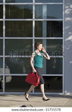 Young woman runs along building while talking on a cell phone. She is wearing a tank top and carrying a red bag. Vertically framed photo.