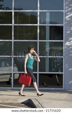 Young woman walks along building while talking on a cell phone and carrying a red bag. She is wearing a tank top. Vertically framed photo.