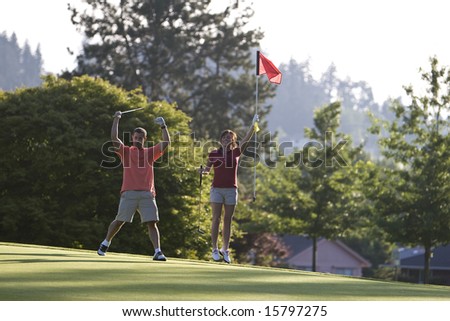 A young couple is playing on a golf course.  The man is holding his golf club above his head and the woman is holding the flag.  They are smiling at the camera.  Horizontally framed shot.