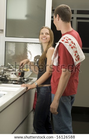 Man and woman stand in kitchen smiling at each other. Woman stirs and man has towel over shoulder. Vertically framed photo.
