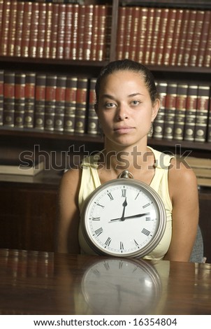 Woman sits at table in a library. She has a clock in front of her resting on table. Vertically framed photo.