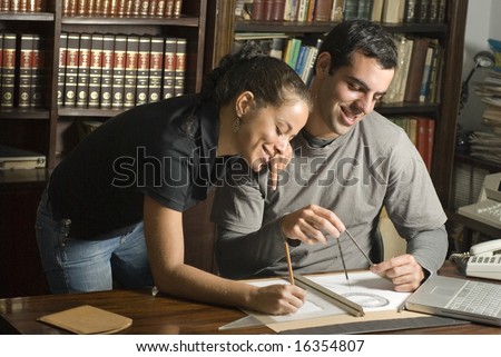 Man sits and woman leans next to him over table. There are maps and tools on the table and they are smiling. Horizontally framed photo.
