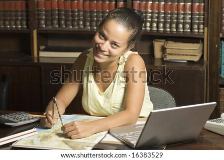 Smiling woman measures map with compass. She is sitting at a desk with a map, laptop, and calculator. Horizontally framed photo.