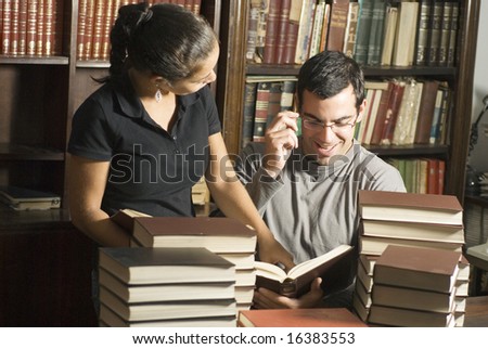 Woman stands while man sits in library. They are reading a book with stacks of books on table. Horizontally framed photo.