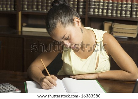 Woman seated at table in library writes in notebook. Horizontally framed photo.