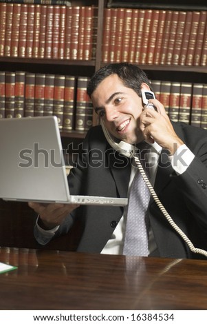 Man in suit sits at a desk while talking on the phone. He is holding a laptop and there are books behind him. Vertically framed photo.