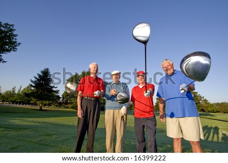 Four elderly men are standing together on a golf course. They are holding out their clubs, smiling, and looking at the camera.  Horizontally framed shot.