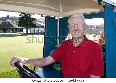 An elderly man is sitting in a golf cart on a golf course.  He is smiling at the camera.  Horizontally framed shot.