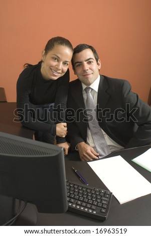 Businessman and woman smile with their heads touching as they work at a desk with a computer. Vertically framed photograph.