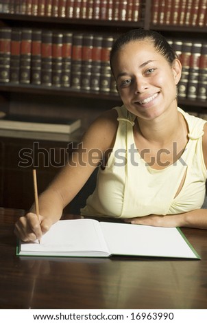 Smiling student working on paperwork while seated at a desk in an office surrounded by books. Vertically framed photo.